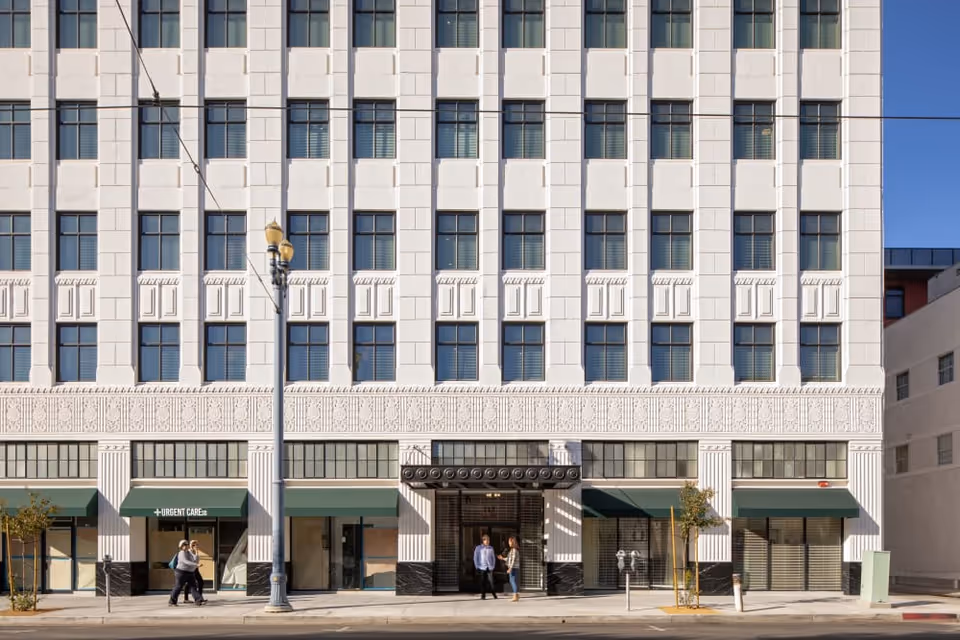 Street-facing facade of a multi-story white historic building with rows of windows, green awnings, a central entrance, and pedestrians on the sidewalk.