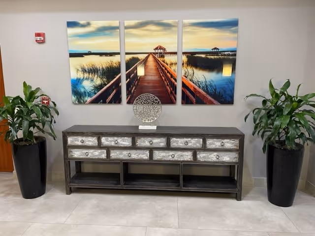 Rustic gray console with multiple drawers flanked by tall potted plants beneath seaside three-panel art.