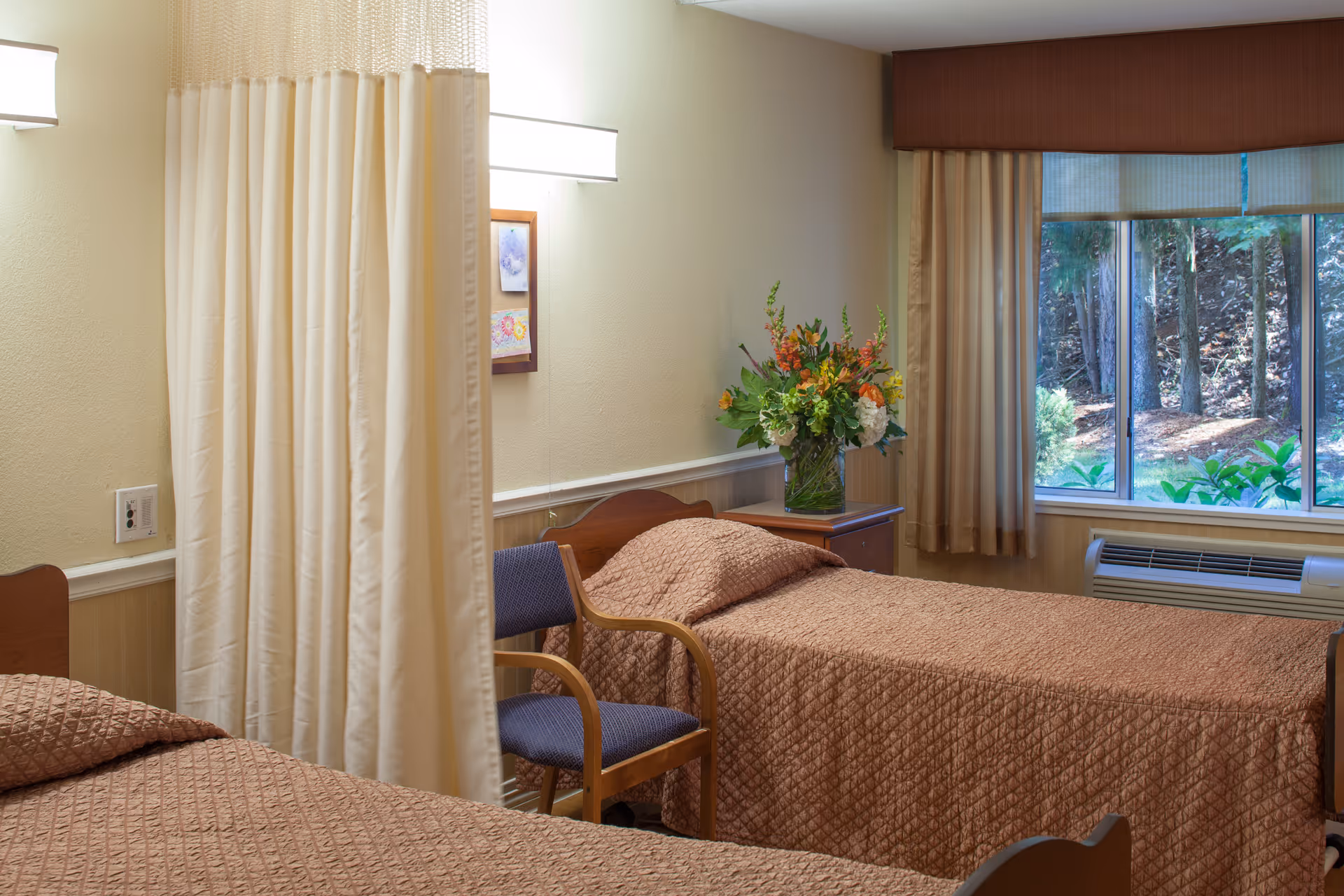 A shared bedroom in a senior living facility with two single beds separated by a privacy curtain. Each bed has a brown quilted bedspread. There is a wooden chair with blue upholstery between the beds, and a nightstand with a vase of colorful flowers. A large window with beige curtains shows a view of trees outside.