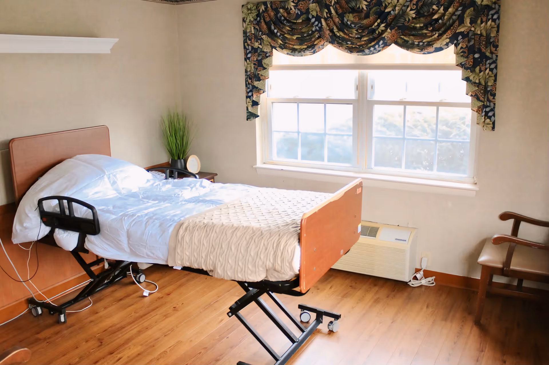 Hospital-style bedroom with adjustable bed, wooden floor, window, and floral curtains.