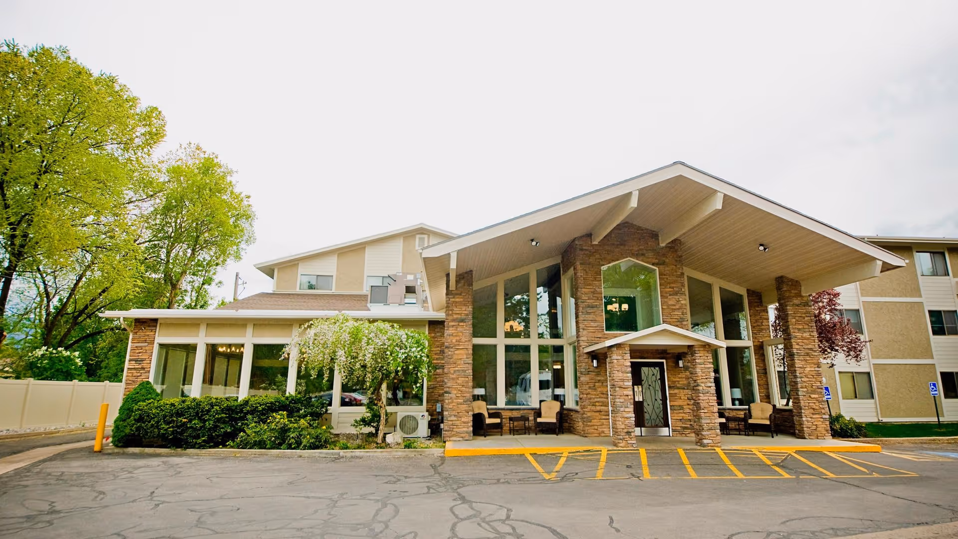 Front exterior view of Williamsburg Retirement Community building with large windows, stone pillars, and a covered entrance. There are trees and shrubs around the building and a parking area with marked spaces in front.