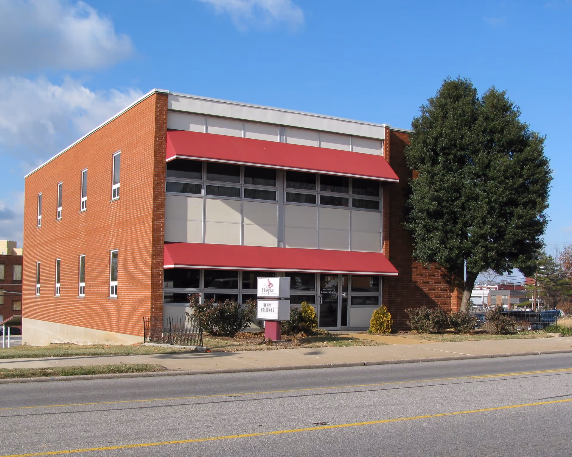Brick hospice facility front with large windows and red awnings along a street.