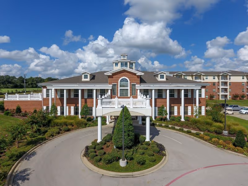 Front exterior of a two-story brick senior living building with a covered porte-cochère, circular driveway, and landscaped roundabout under a blue sky.