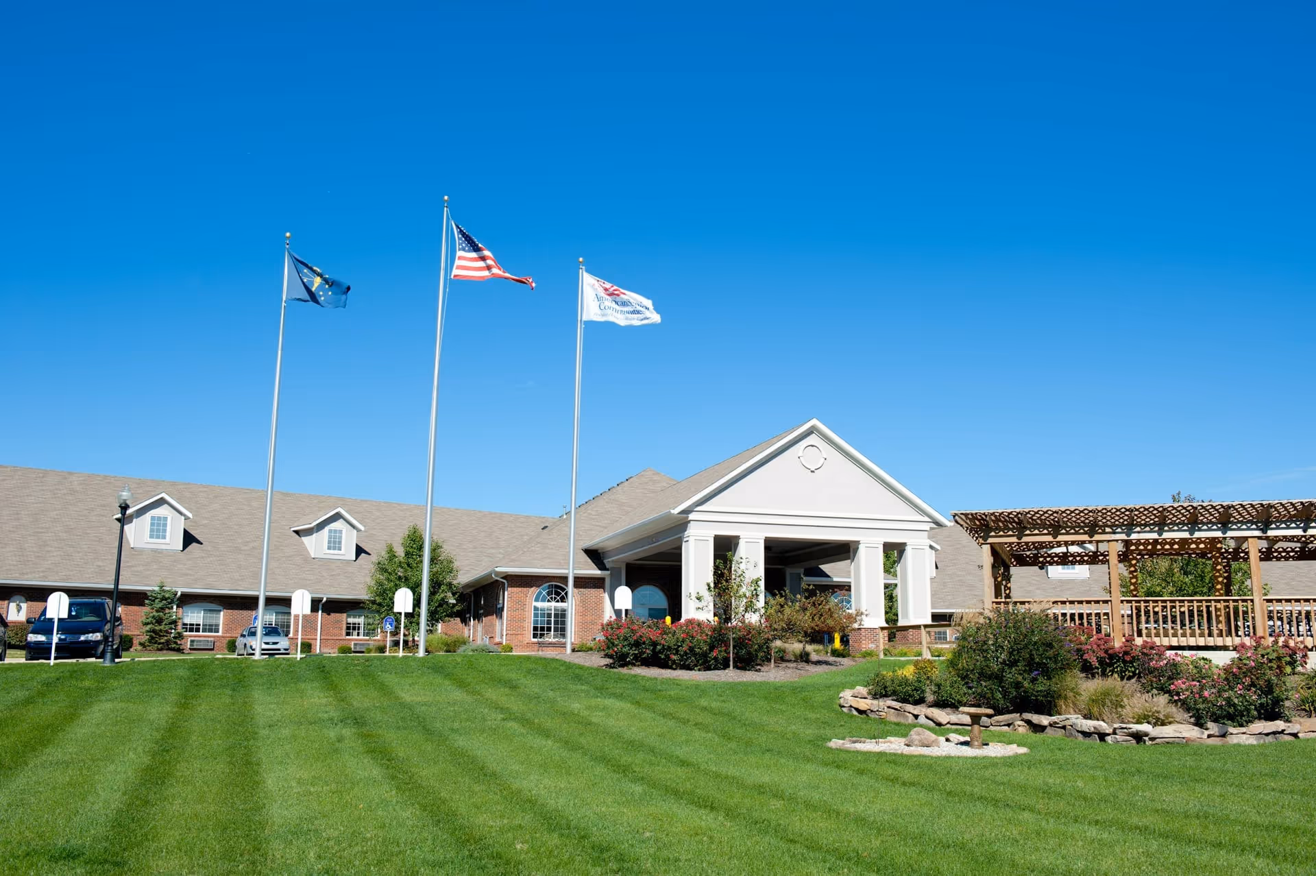 Front exterior of Coventry Meadows with flagpoles, a covered entrance, manicured lawn and a wooden pergola.