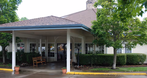 Entrance of a senior living facility with a covered driveway, a wooden bench, potted plants, and surrounding greenery including trees and bushes.