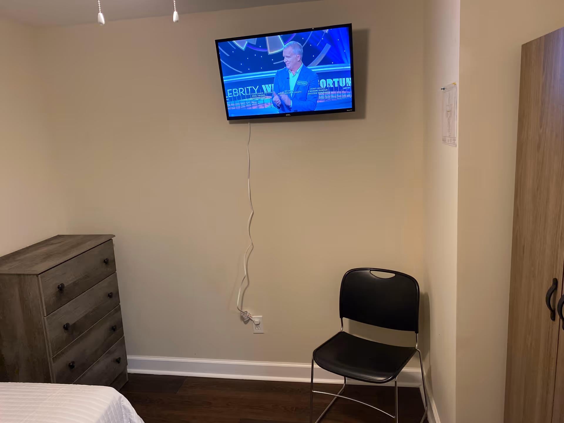 Small bedroom with dresser, bed edge, wall-mounted TV, black chair, beige walls.