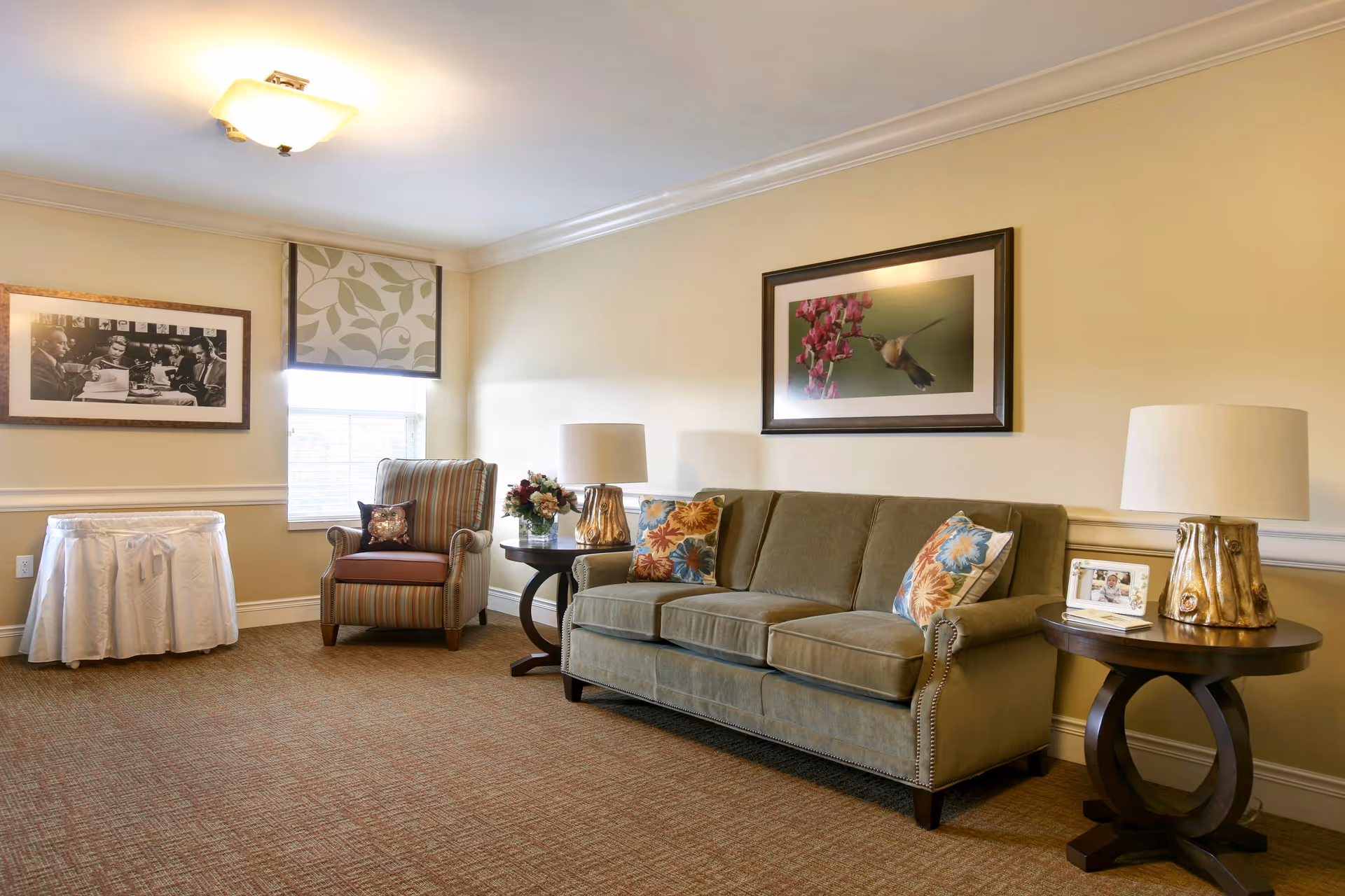 Cozy senior living common area with sofa, armchair, tables, lamps, and wall art.