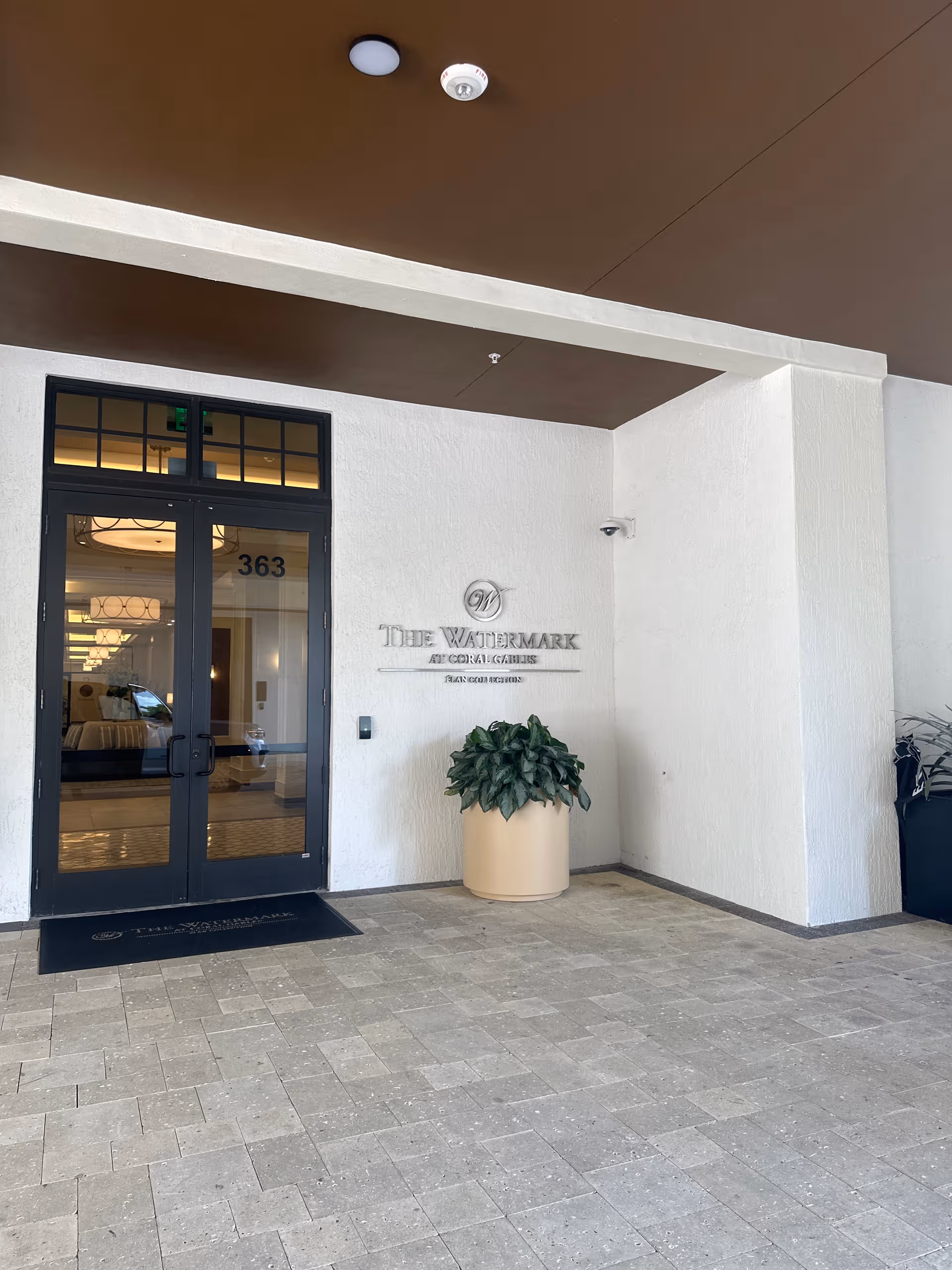 Exterior entrance of a senior living facility with glass doors and a tall potted plant.