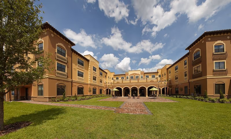 Courtyard view of a three-story Mediterranean-style senior living building with a green lawn, brick pathways and shaded patio seating under a partly cloudy sky.