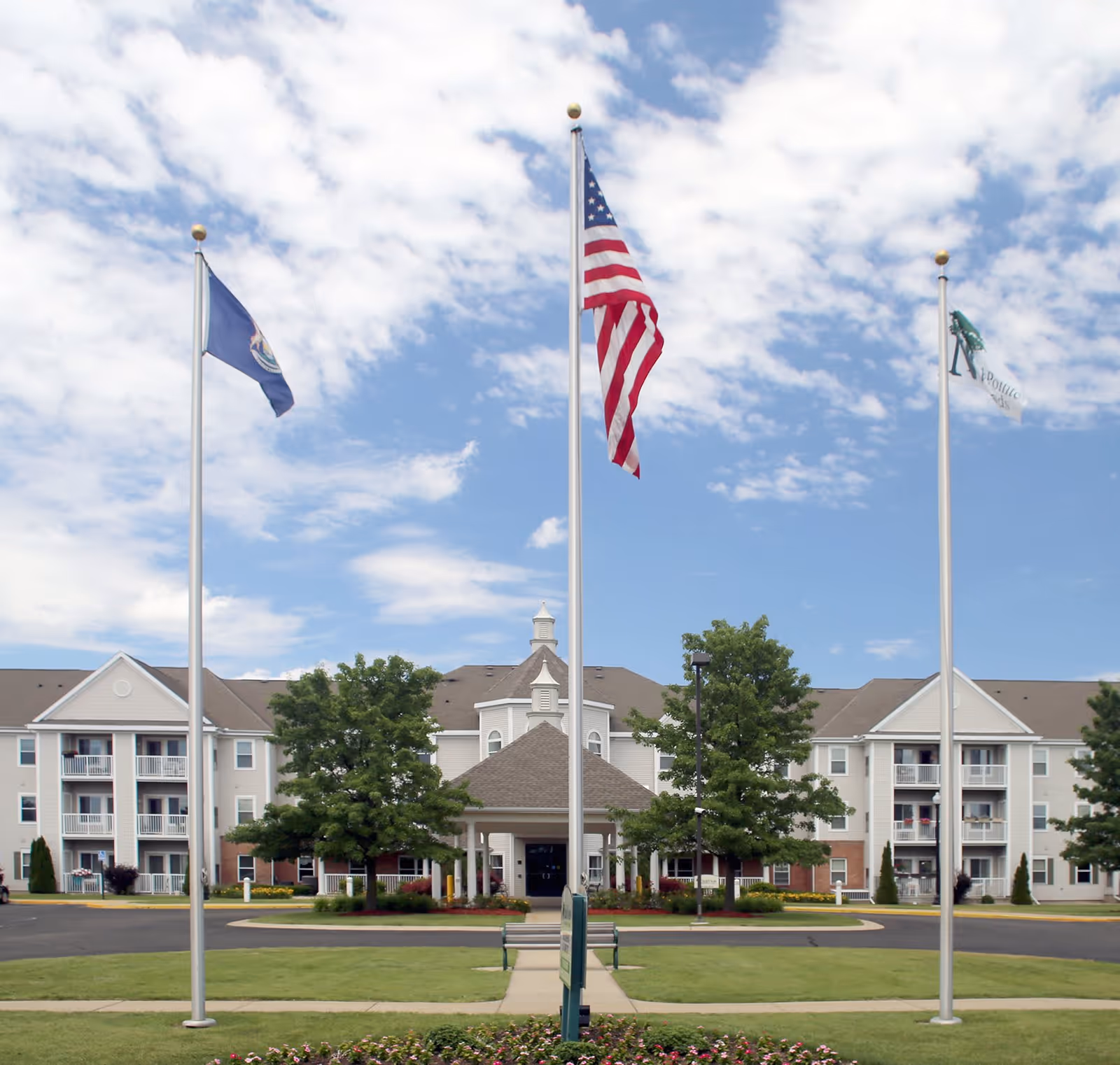 Exterior view of NorthPointe Woods senior living facility with three flagpoles in front displaying the American flag, a state flag, and a facility flag. The building is a multi-story structure with balconies, surrounded by trees and a well-maintained lawn under a partly cloudy sky.