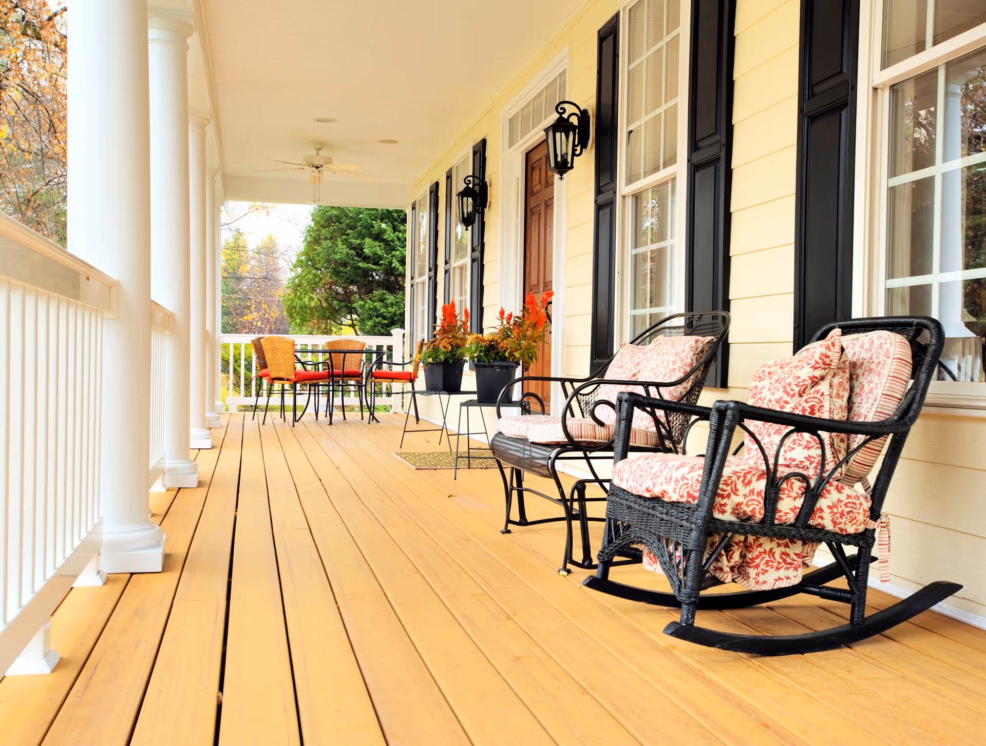 Sunny front porch with wicker chairs, potted plants, and a table.