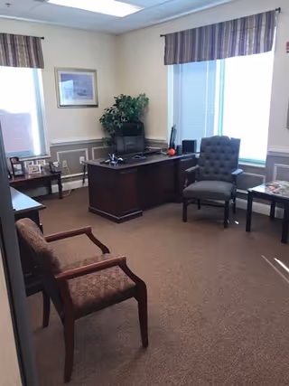 A small, well lit office with a desk, chairs, plants, and windows.