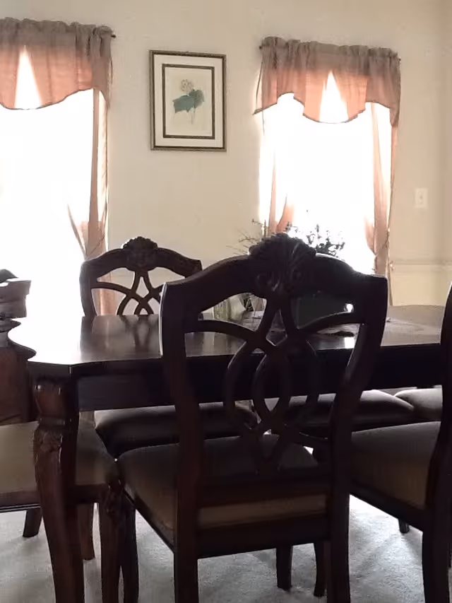 Dark wood dining table with ornate chairs in a room with two curtained windows and a framed picture on the wall.