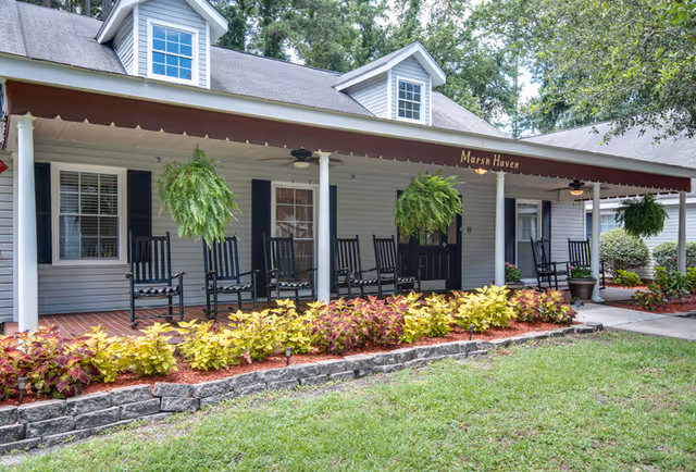 Front porch of a single-story building with a brown awning labeled 'Marsh Haven'. The porch features several black rocking chairs, hanging green ferns, and a landscaped garden with colorful plants bordered by stone edging. The building has gray siding, white columns, and dormer windows on the roof.