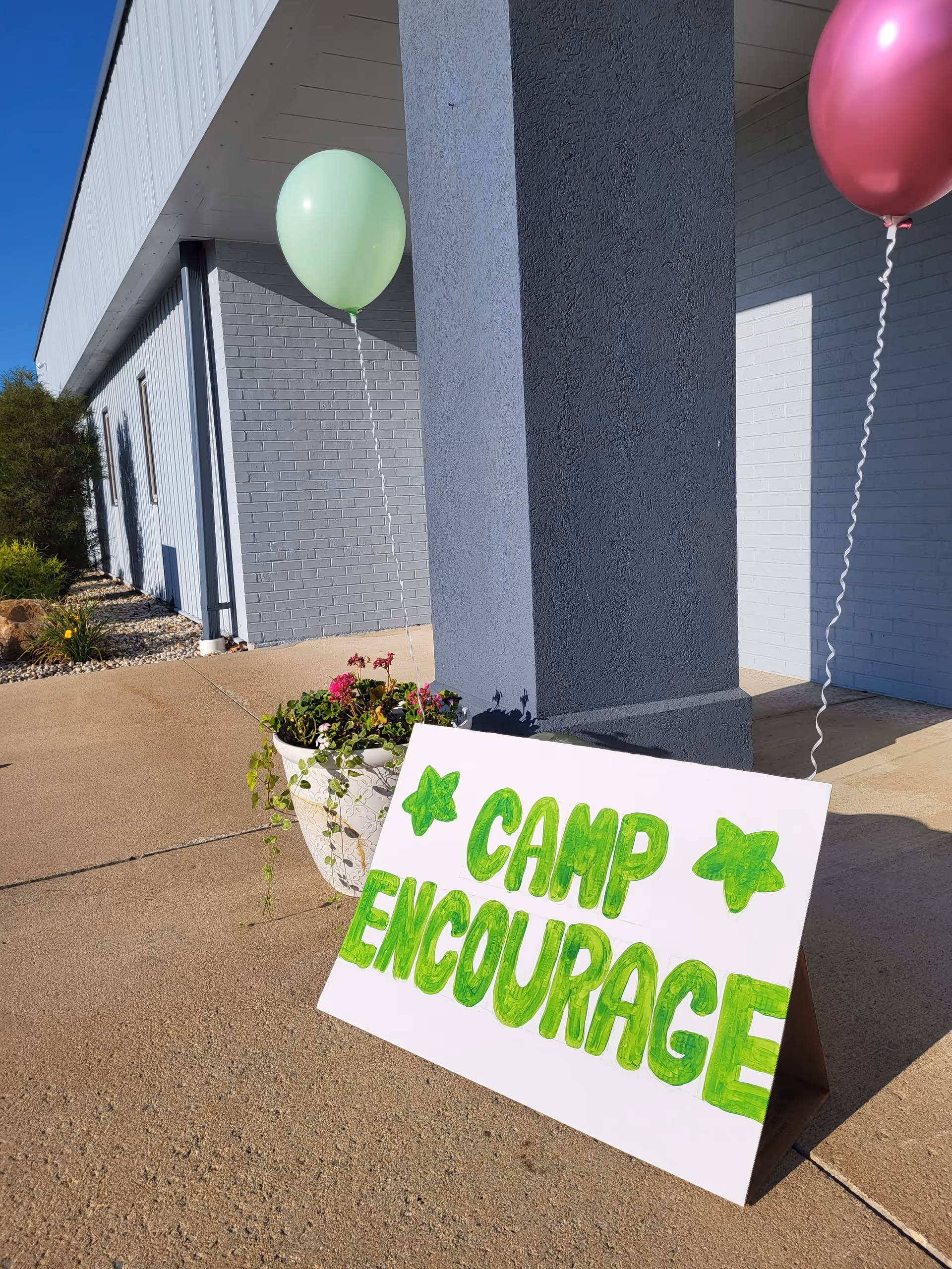 Exterior of a facility shows balloons, planter, and a sign reading Camp Encourage near a pillar.