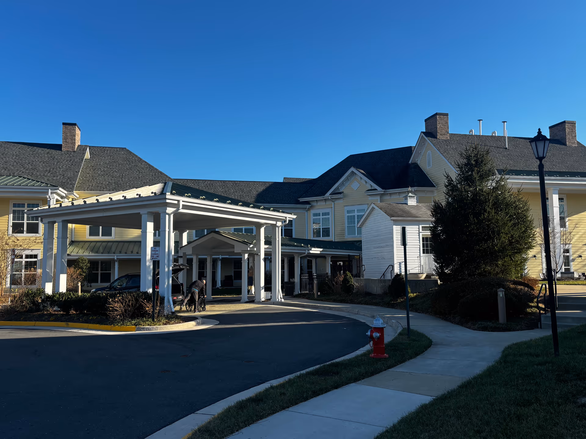 Covered entrance (porte-cochere) of a yellow senior living building with a driveway and clear blue sky.