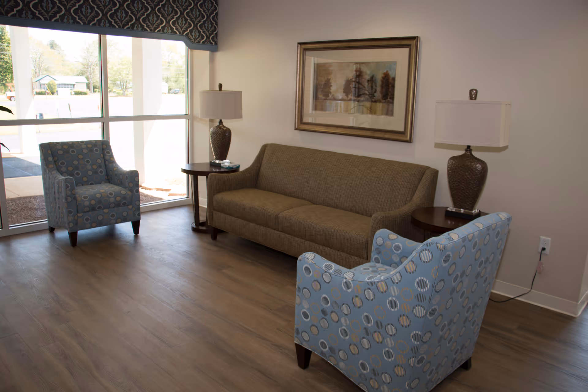 A cozy senior living lounge with blue patterned chairs, a tan sofa, and lamps.