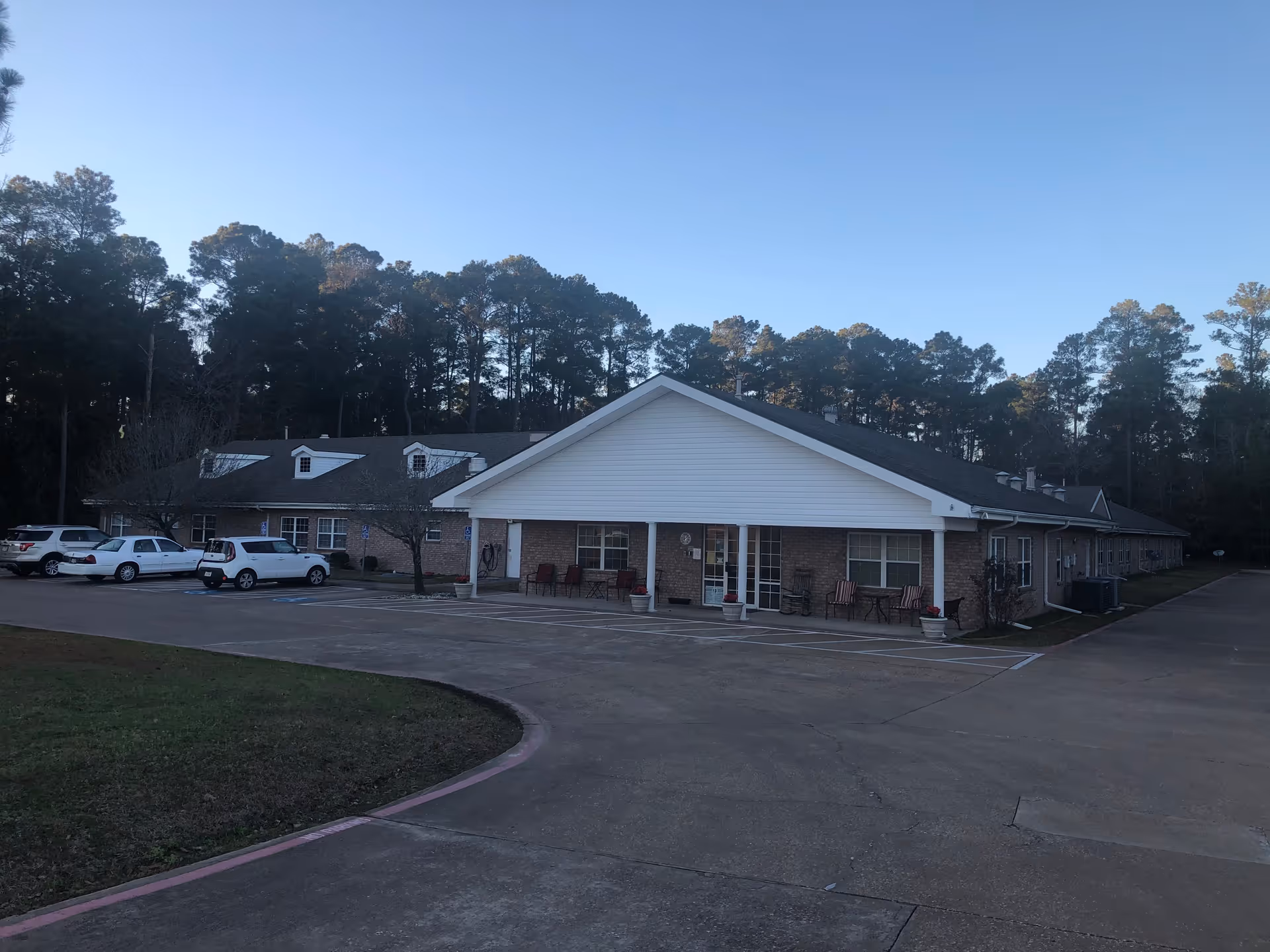 Exterior view of a single-story senior living facility building with a brick facade and white siding on the front gable. Several cars are parked in the parking lot in front of the building. The building is surrounded by tall trees under a clear blue sky.