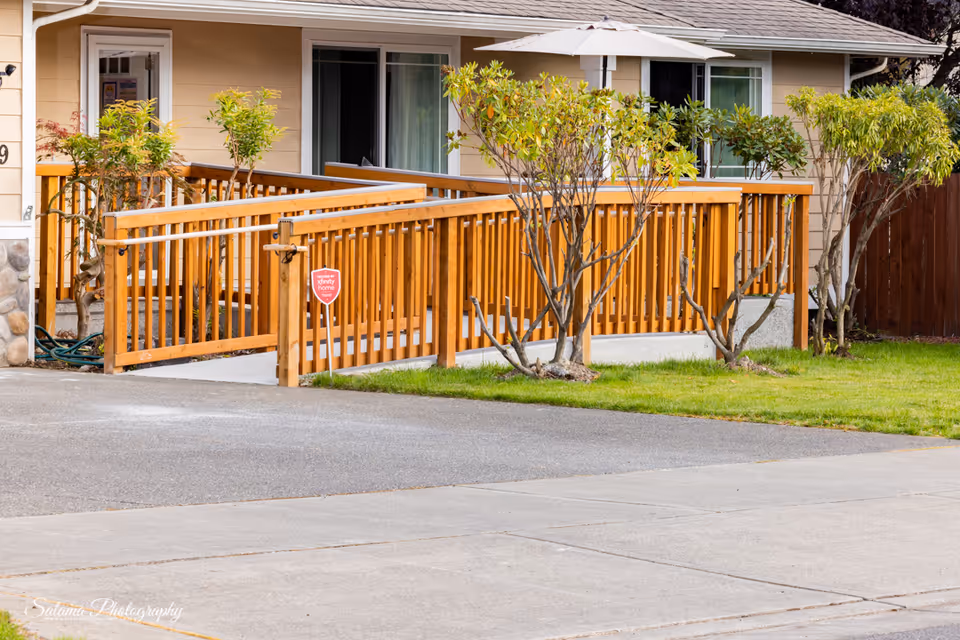 Exterior view of a senior home with a wooden ramp, deck railing, and nearby trees.