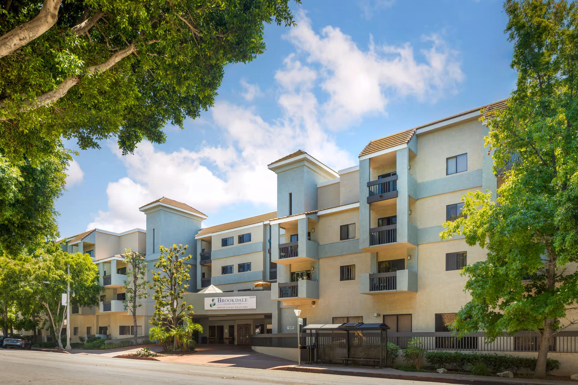 Exterior view of Brookdale Uptown Whittier senior living facility, a multi-story building with balconies, surrounded by trees under a partly cloudy sky.