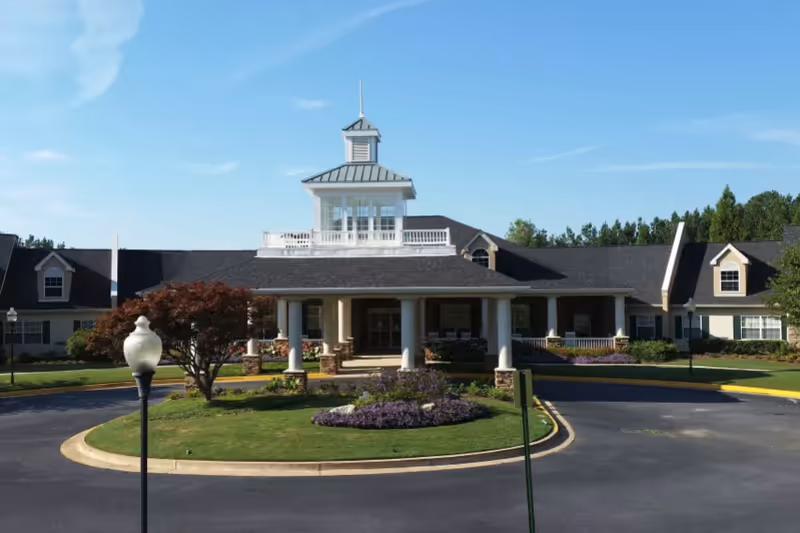 Front exterior view of a large senior living facility building with a covered entrance supported by white columns, a circular driveway, landscaped greenery, and a clear blue sky.