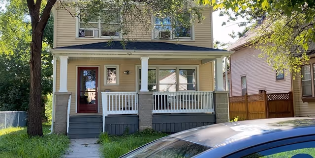 Two-story beige house with a covered front porch and white railings on a sunny day.