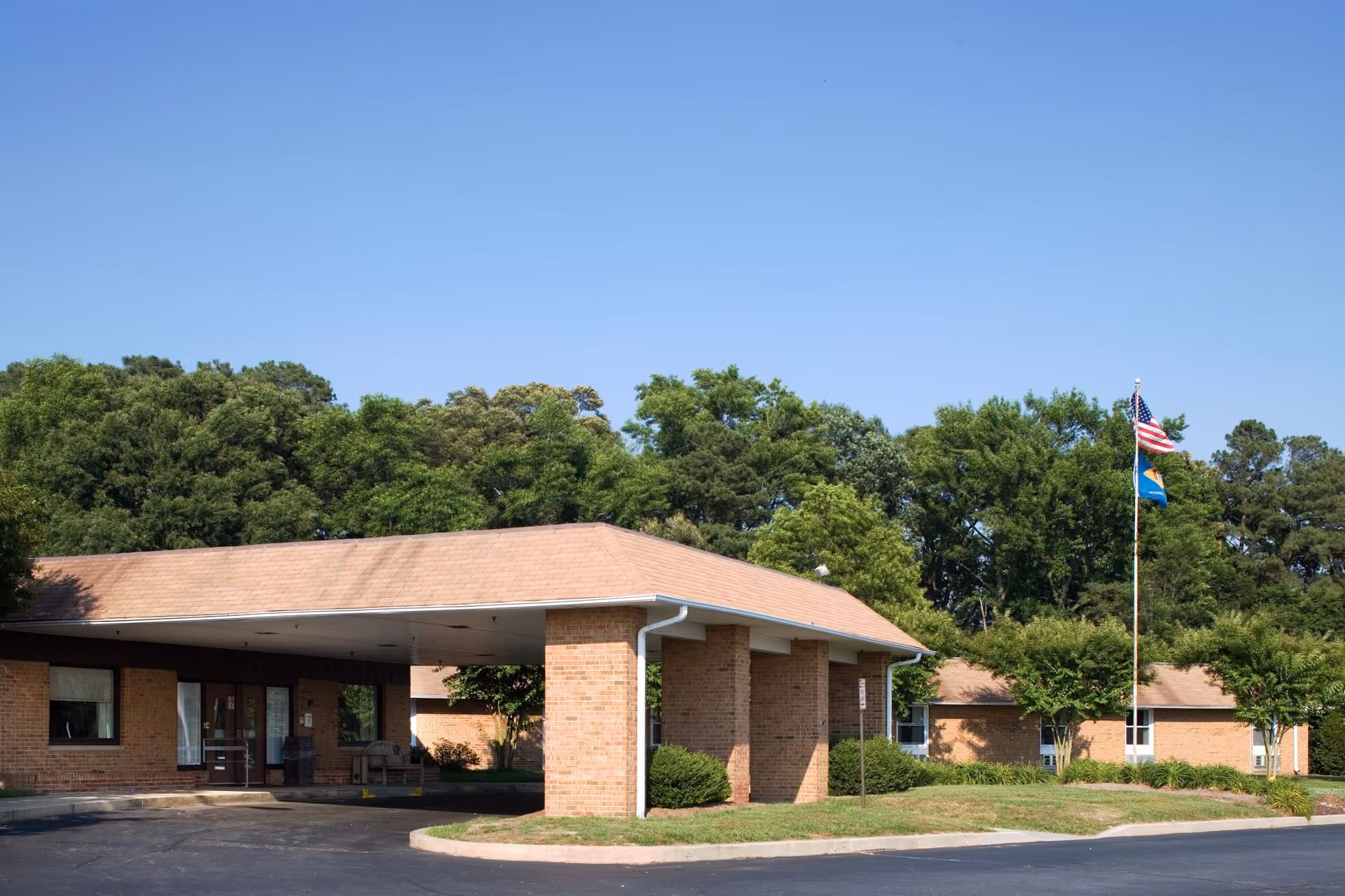 Exterior view of a single-story brick building with a covered entrance, surrounded by green trees and bushes under a clear blue sky. Two flags, including an American flag, are flying on a flagpole near the building.