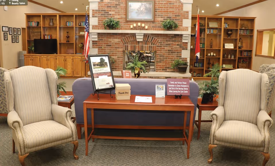 A cozy common area with two striped armchairs facing a wooden table that holds informational signs and a thank you box. Behind the table is a purple couch, and in the background, there is a brick fireplace flanked by wooden bookshelves filled with books and decorative items. An American flag and another flag are positioned on either side of the fireplace.