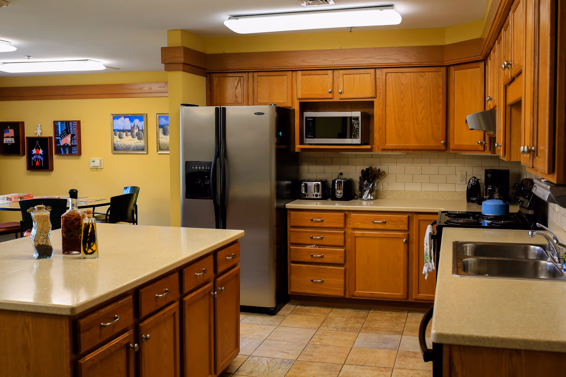 A kitchen area with wooden cabinets, a stainless steel refrigerator, microwave, toaster, coffee maker, and a stove with a blue kettle. There is a large island countertop in the foreground with jars containing food items. In the background, a dining area with tables and chairs is visible, along with framed pictures on the yellow walls.