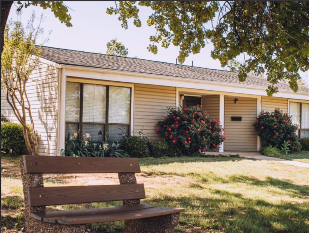 A single-story residential building with beige siding and a shingled roof, surrounded by greenery and flowering bushes. In the foreground, there is a wooden bench on a grassy area under the shade of a tree.