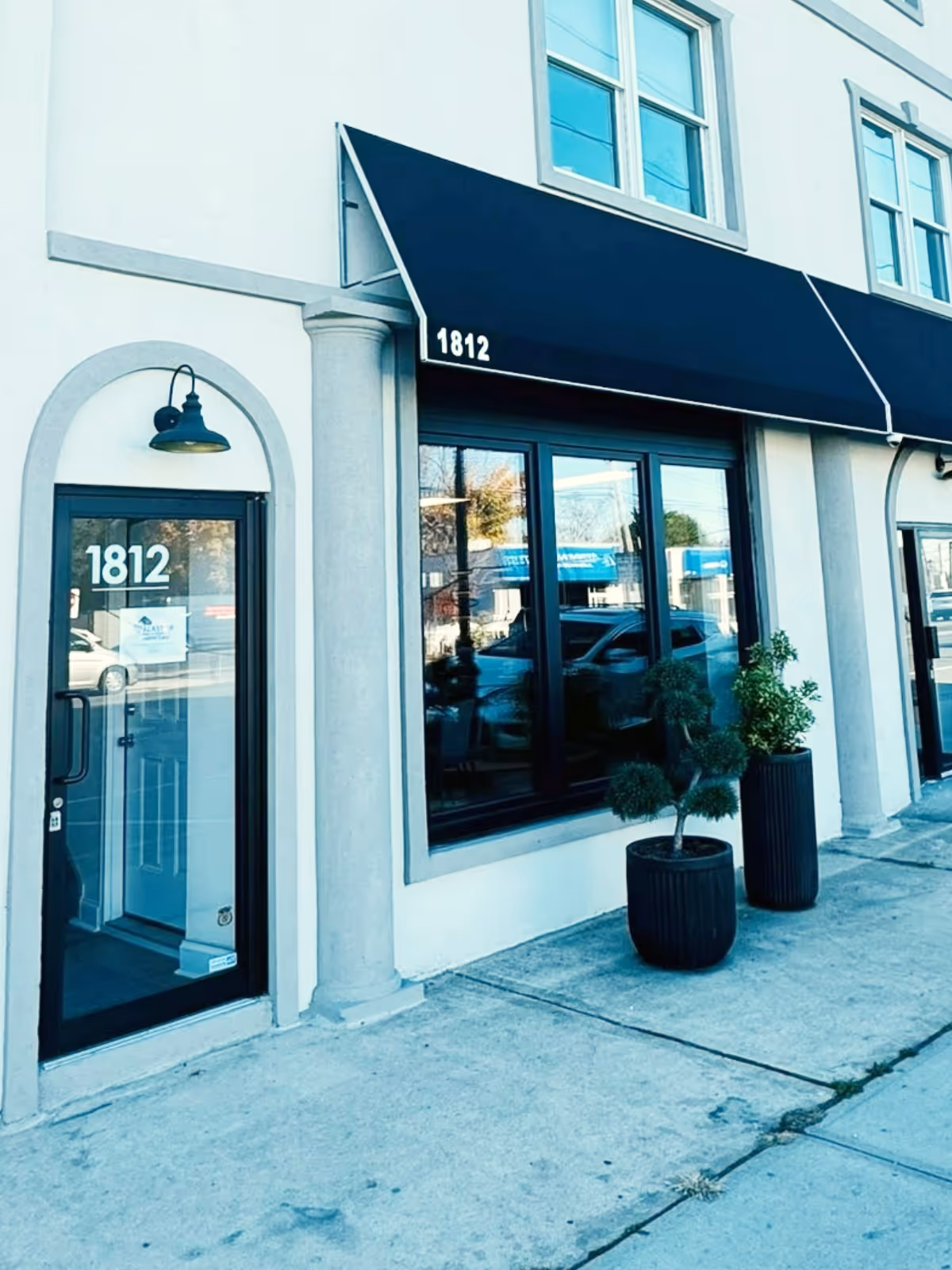 Storefront with black awning, glass doors and windows, address 1812, potted plants outside.