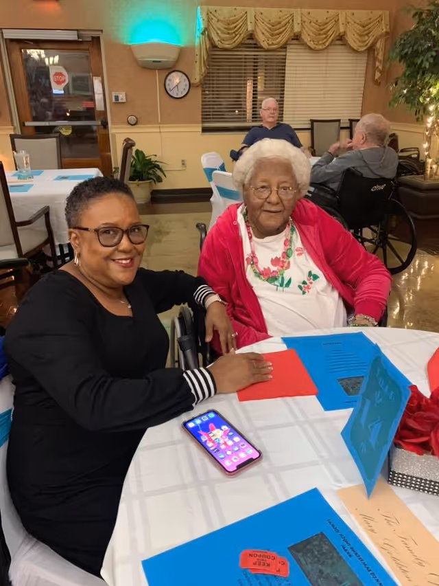 Two women sitting at a round table in a dining area of a senior living facility. One woman is wearing glasses and a black top, smiling at the camera. The other woman, with white hair and glasses, is wearing a white shirt with floral print and a red jacket. There are blue and red papers on the table, along with a smartphone. In the background, two elderly men are seated, one in a wheelchair. The room has beige walls, a window with blinds and a valance, a clock, and some plants.