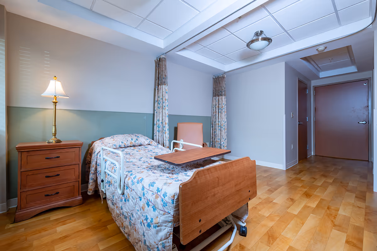 A patient bedroom in a facility featuring a bed, dresser, lamp, and curtains.