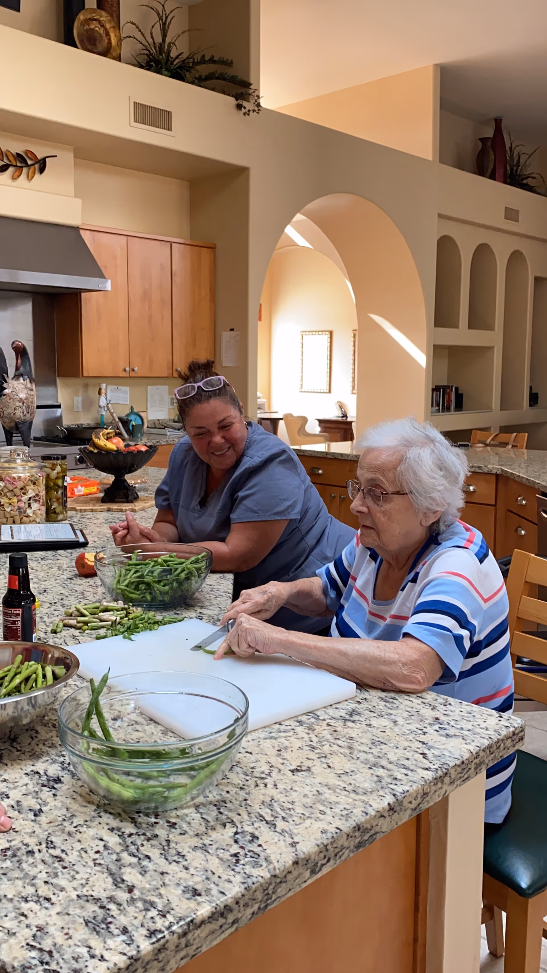 An elderly woman and a caregiver are sitting at a kitchen counter preparing green beans. The elderly woman is cutting the beans on a white cutting board while the caregiver watches and smiles. The kitchen has wooden cabinets, a granite countertop, and various kitchen items including bowls of green beans and a fruit bowl.