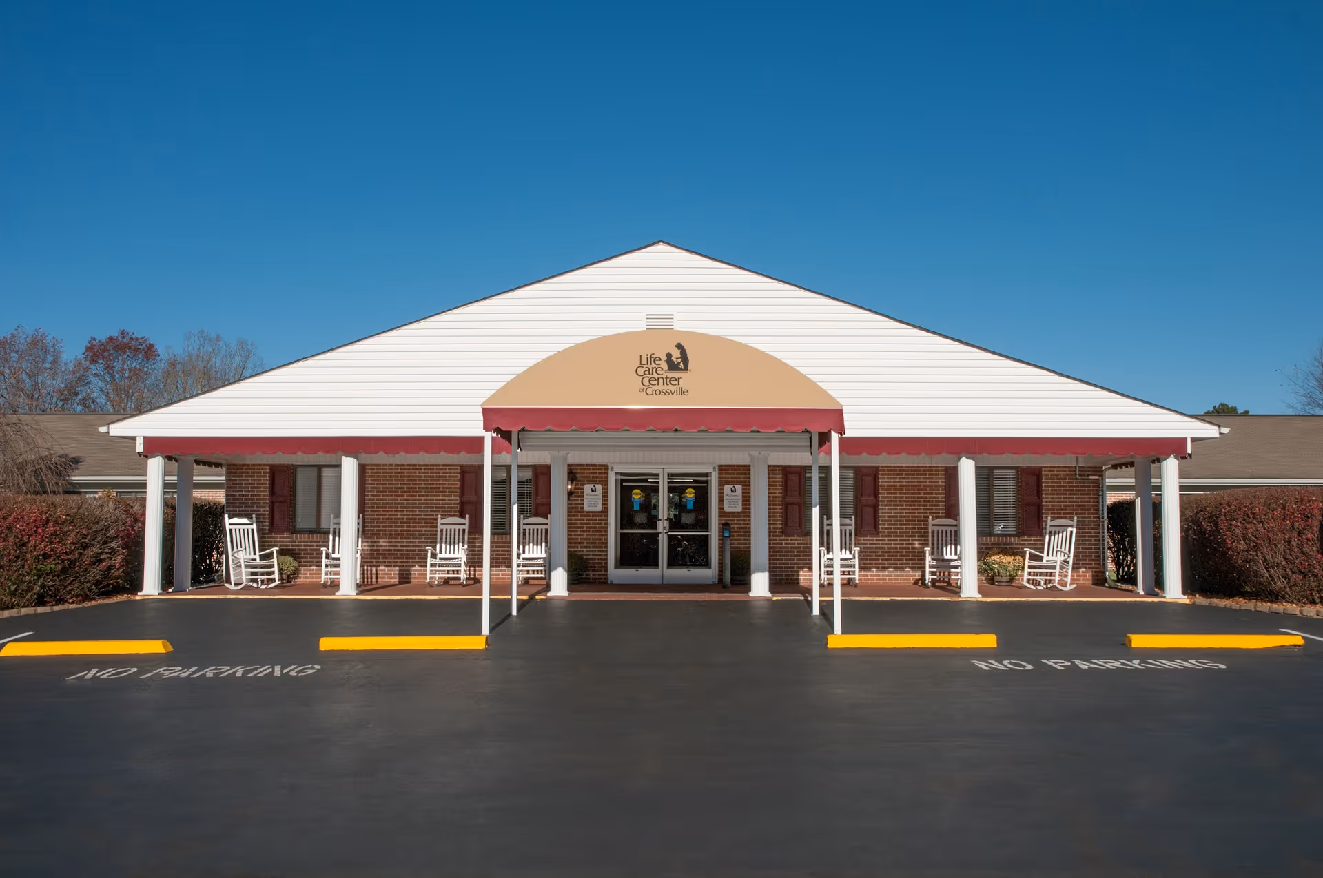 Front exterior view of the Life Care Center of Crossville building with a covered entrance, white columns, several white rocking chairs on the porch, and clear blue sky above.