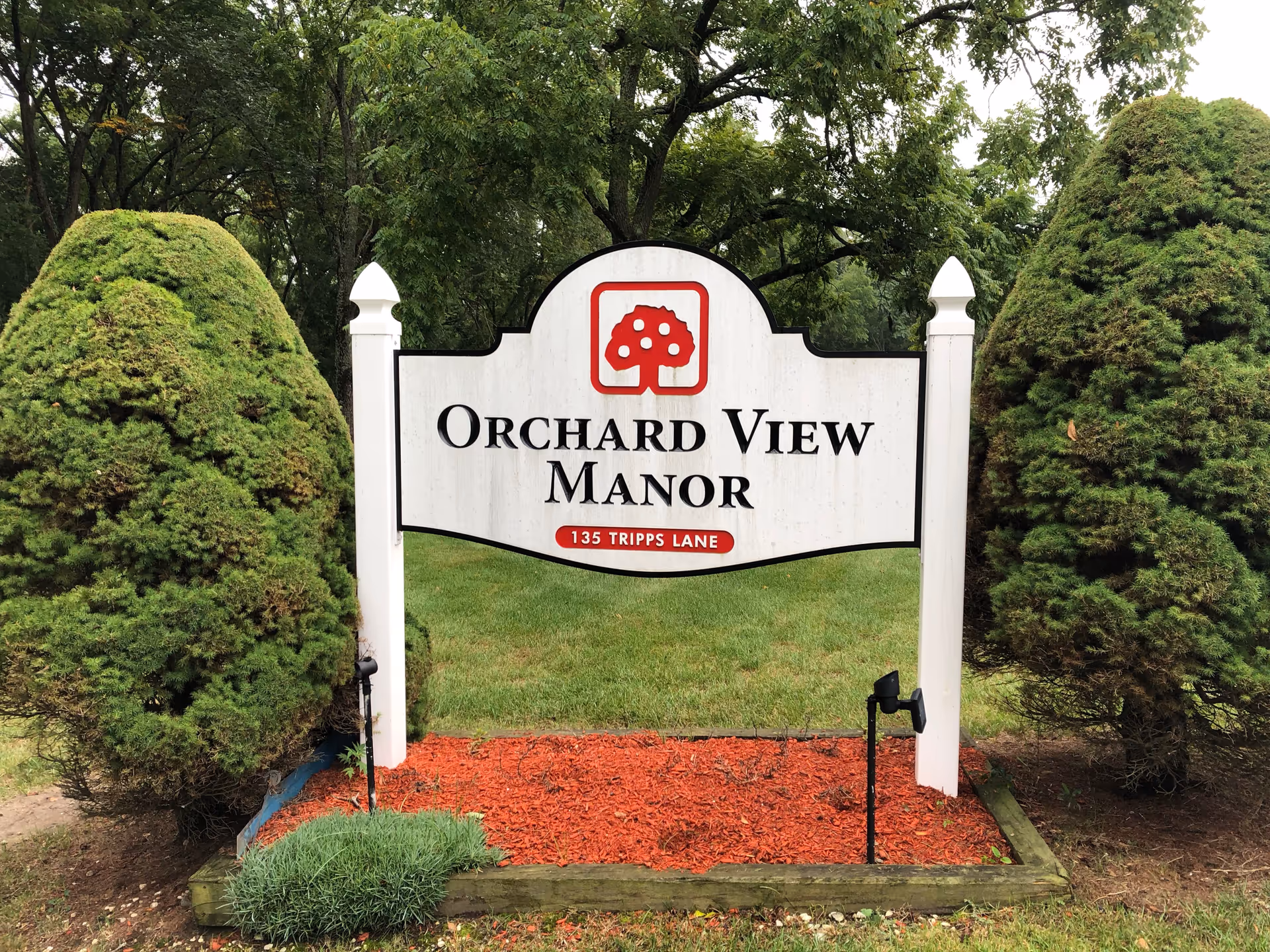 White wooden sign reading "Orchard View Manor" surrounded by trimmed bushes, mulch, and trees in the background.