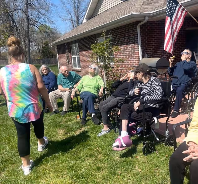 Senior residents sit and chat in a sunny outdoor seating area by a brick facility.