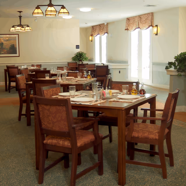 A dining room in a senior living facility with wooden tables and chairs arranged neatly. Each table is set with plates, glasses, napkins, and condiments. The room is well-lit with ceiling lights and natural light coming through large windows with curtains. There are plants and framed artwork on the walls, creating a warm and inviting atmosphere.