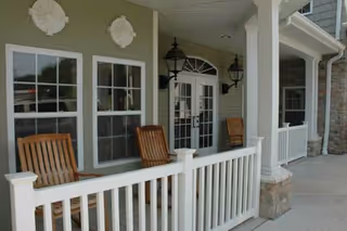 Front porch area of a senior living facility with white railing, two wooden rocking chairs, decorative wall accents, and double glass doors under a covered entrance.