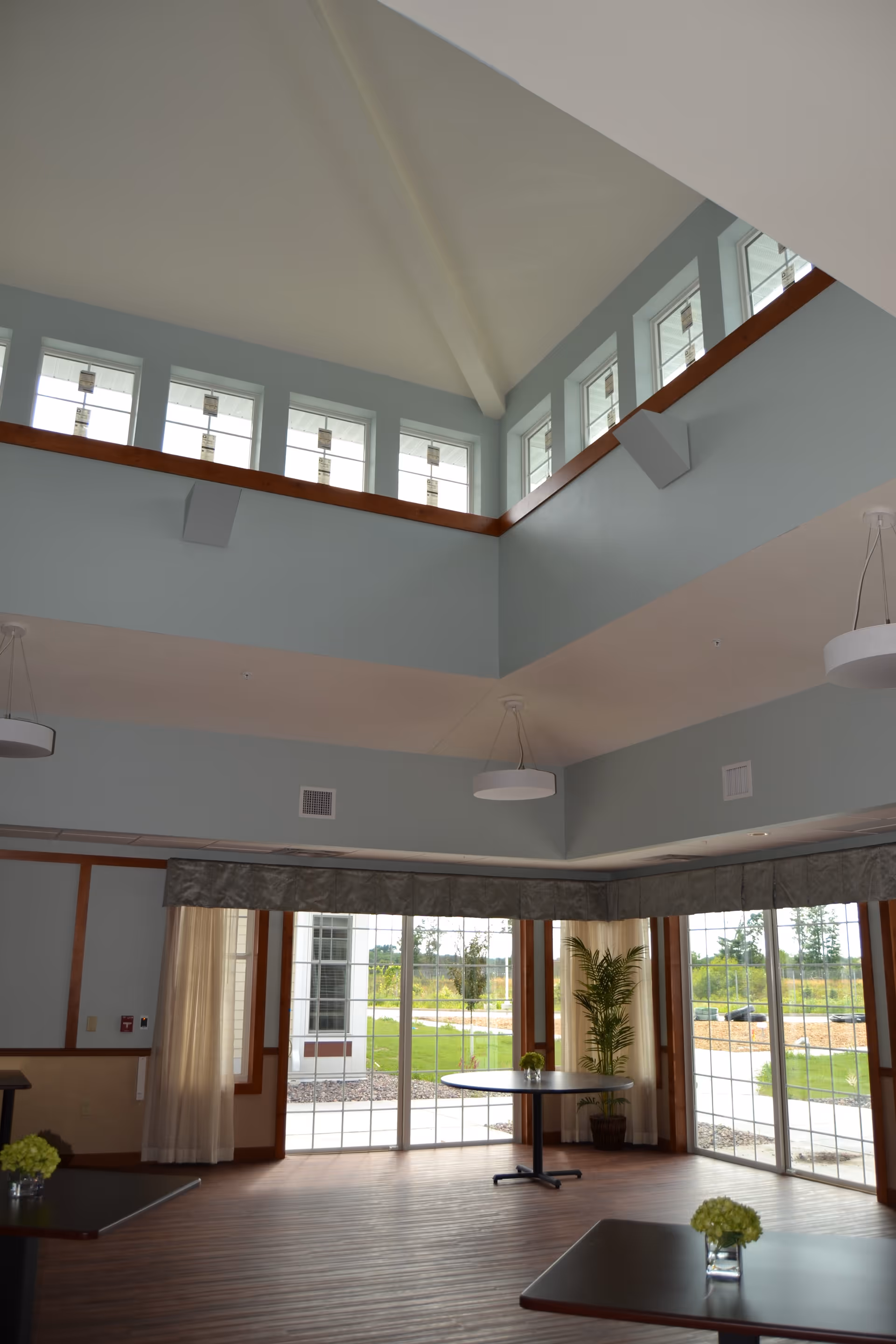 Bright double-height common room with a vaulted ceiling, clerestory windows, pendant lights, tables, and large sliding glass doors to an outdoor area.