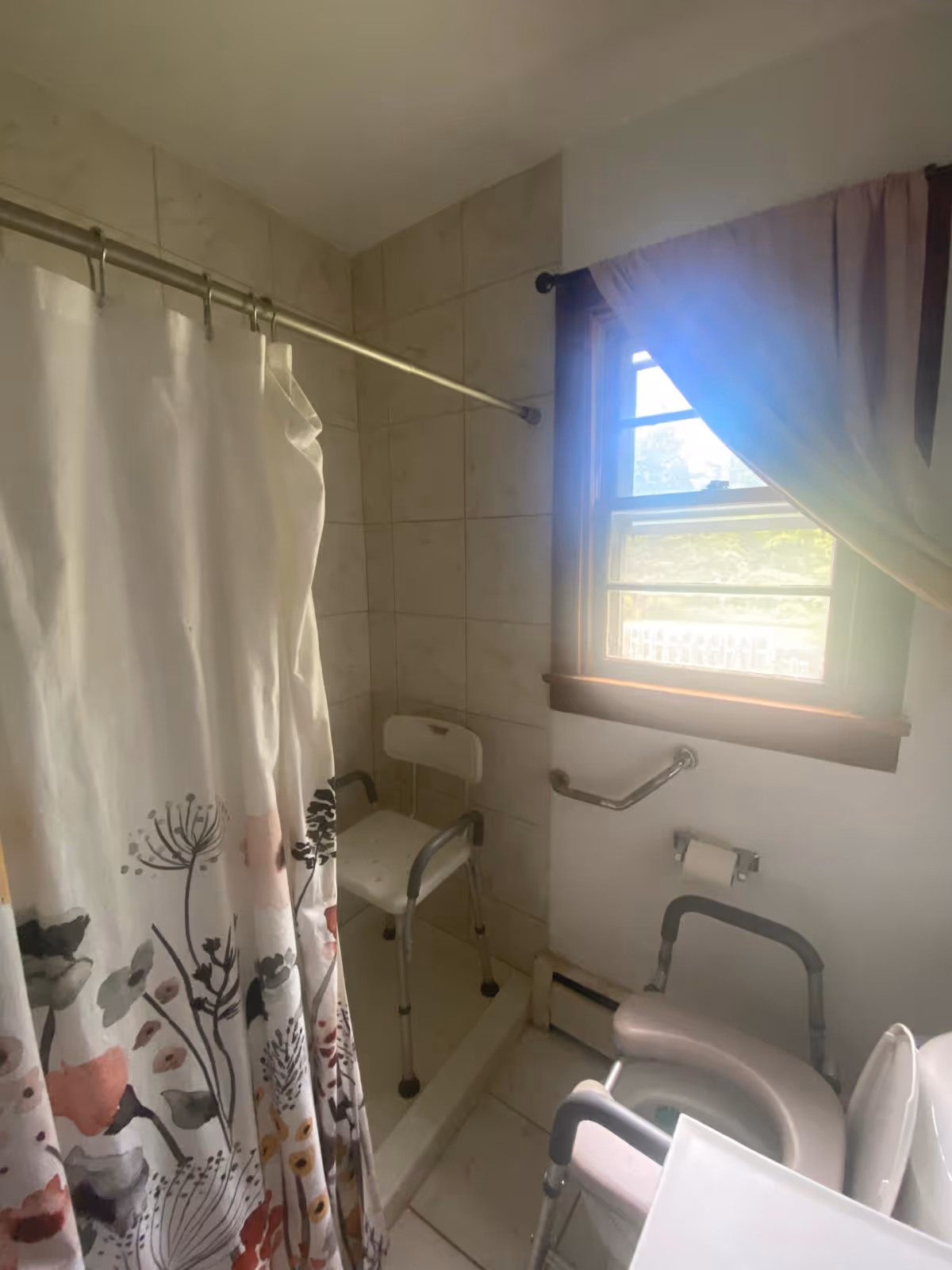 Compact bathroom in a senior living facility with floral shower curtain and chair.