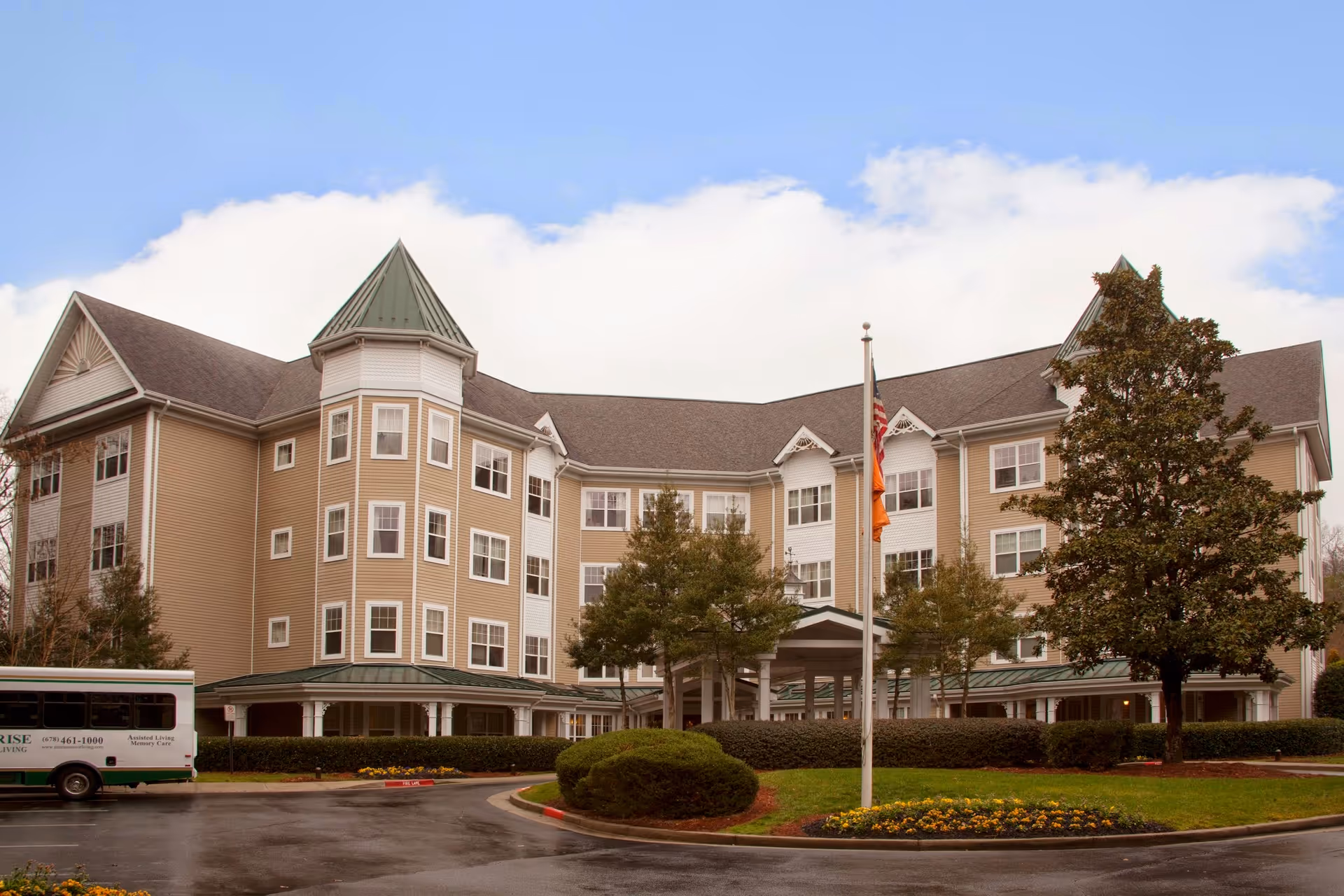 Exterior view of a large, multi-story senior living facility building with beige siding and a green roof. The building has multiple windows and a covered entrance with a driveway in front. There are trees, shrubs, and a flower bed surrounding the driveway, and a flagpole with flags near the entrance. A shuttle bus is parked on the left side of the image.