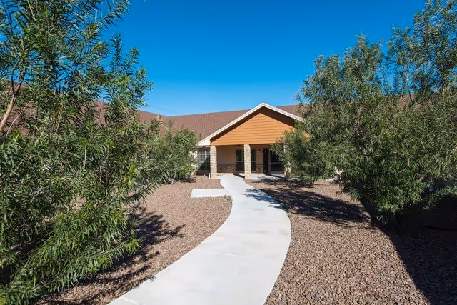 A paved walkway leading through a landscaped area with trees and gravel towards the entrance of a single-story building with a brown roof and a covered porch.