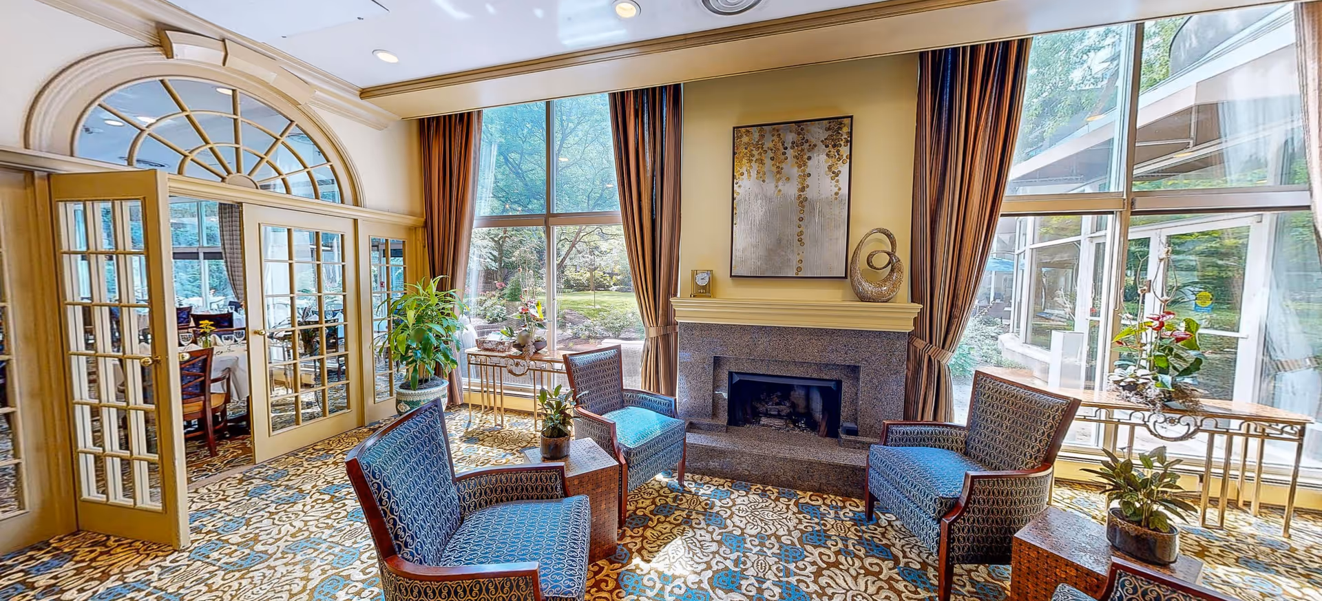 Cozy senior living lounge featuring a fireplace, blue chairs, and large sunlit windows.