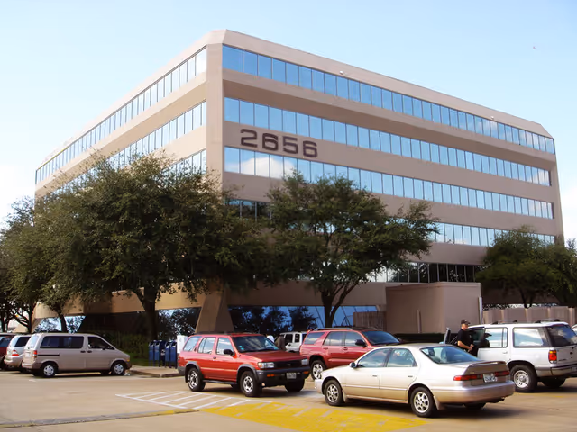 Three-story beige office building with large windows and parked cars in front.