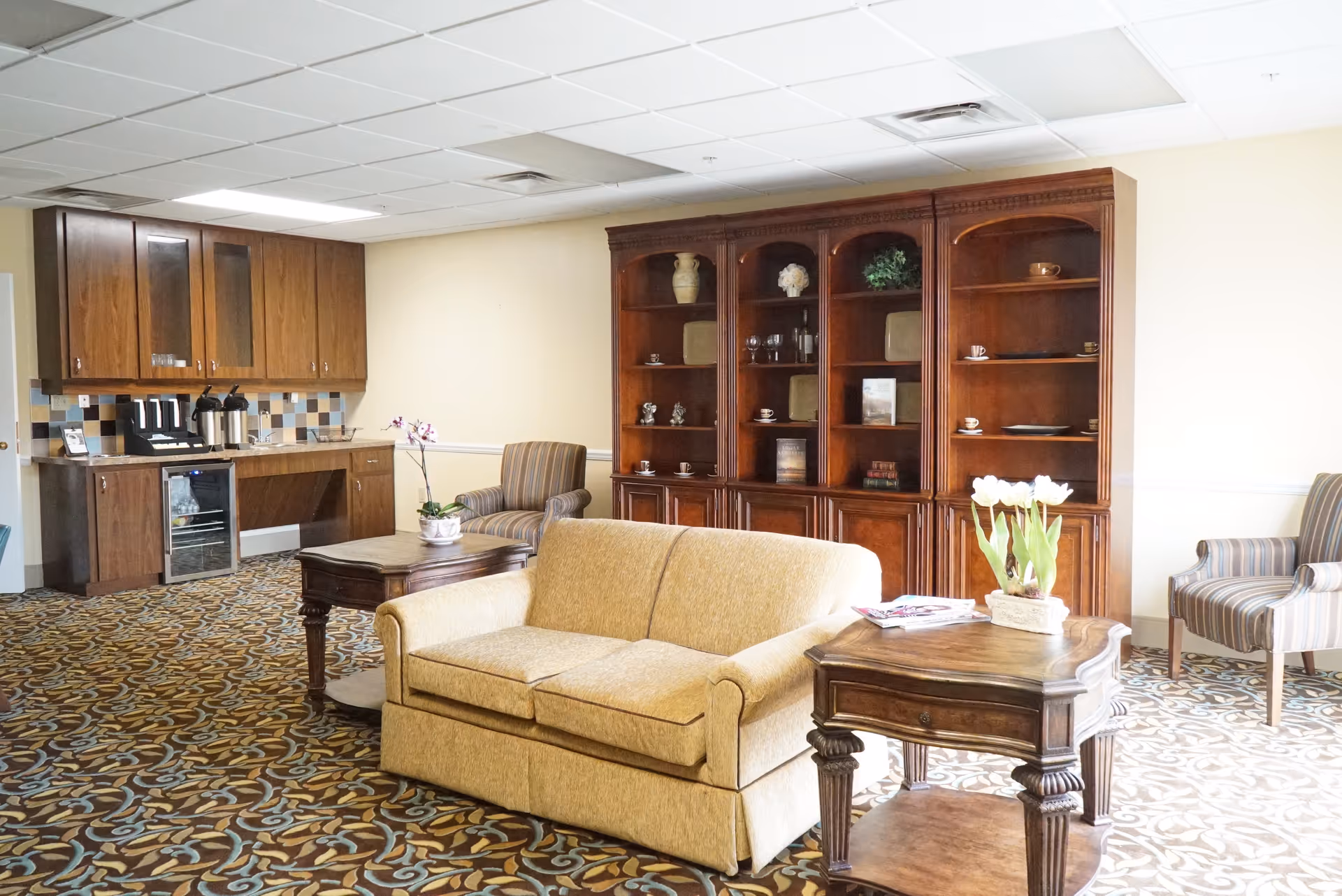 A furnished common room with a sofa, side tables, armchairs, a large wooden shelving unit, and a small kitchenette area.