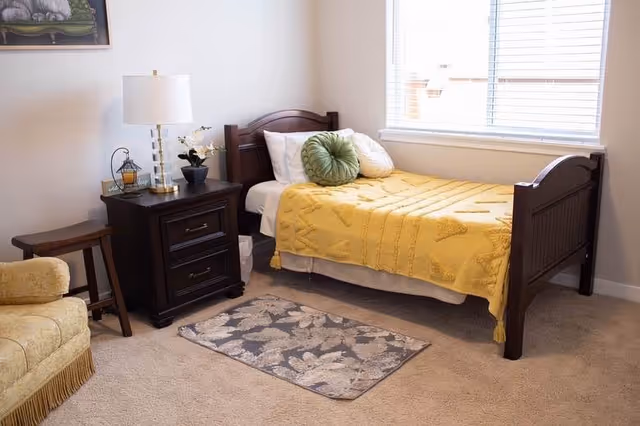 A cozy bedroom with a single wooden bed covered with a yellow blanket and decorative pillows. Next to the bed is a dark wooden nightstand with a lamp, a small plant, and a lantern. There is a small patterned rug on the carpeted floor, a wooden stool, and a beige armchair partially visible on the left side. A window with blinds allows natural light into the room.