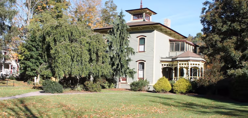 A large, two-story historic house with light green siding and a small tower on the roof, surrounded by trees and bushes with a grassy lawn in front under a clear blue sky.