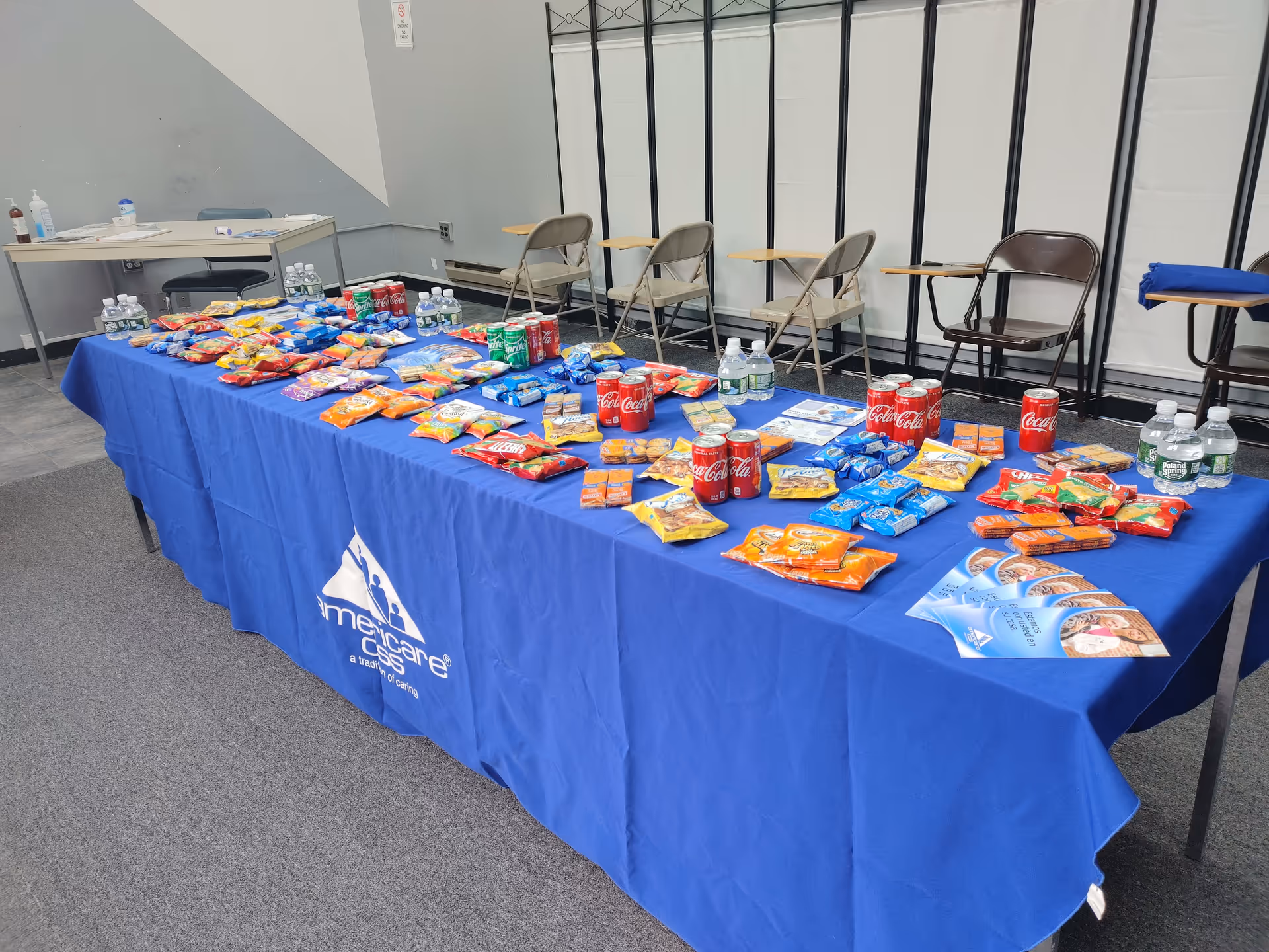 Long table covered with blue cloth at Americare Coleman Street, loaded with snacks and water bottles.