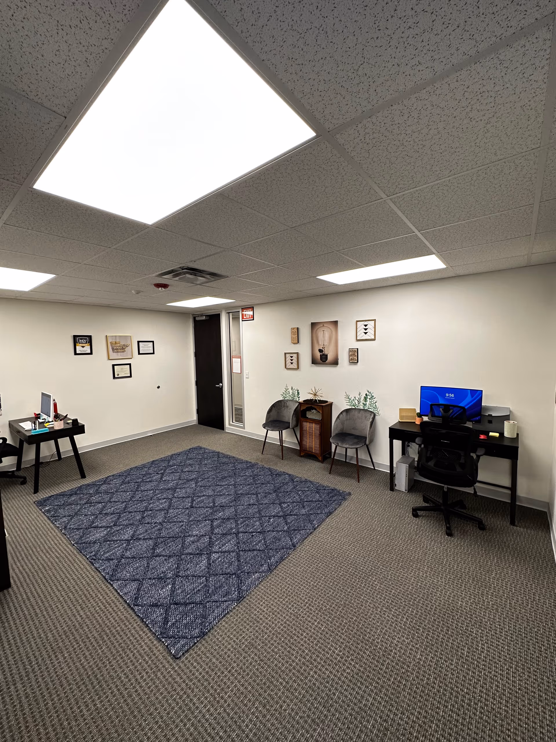 Interior office area with desks, computer, chairs, framed art, and a blue rug.